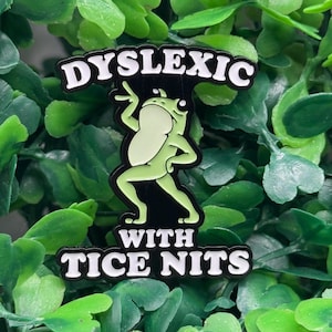 May include: A black enamel pin with the words "Dyslexic" above and "With Tice Nits" below a green frog illustration. The frog is in a dancing pose with its arms and legs outstretched. The pin is surrounded by green leaves.