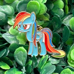 Op de afbeelding: Een blauwe emaille pin met een cartoon pony met een regenboog manen en staart. De pony heeft een gouden omtrek en is geplaatst tegen een achtergrond van groen gebladerte. De pin is een verzamelobject.