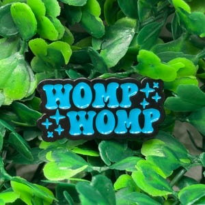 May include: A black enamel pin with the text "WOMP WOMP" in light blue bubble letters. The pin also features light blue starburst designs. The pin is set against a background of green leaves.