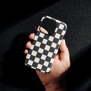 Può includere: Custodia per telefono a scacchi bianchi e neri tenuta in mano. La custodia presenta un motivo ripetuto di quadrati neri e crema. La custodia del telefono è rettangolare con angoli arrotondati e un ritaglio per la fotocamera.