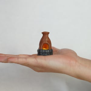May include: A small, brown clay fireplace with a black base and a glowing orange fire inside. The fireplace is sitting on the palm of a hand.