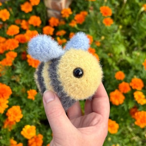 May include: A fuzzy, round, crocheted bee toy featuring a yellow head, a body with alternating dark gray and yellow stripes, and two light blue wings. The bee has a single black plastic bead eye visible on the side of its head.