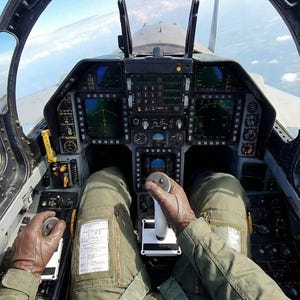 May include: The cockpit of a fighter jet featuring a central control stick, multiple digital display screens, and various toggle switches. The view shows the pilot's legs in olive green flight suit trousers and brown leather gloves.