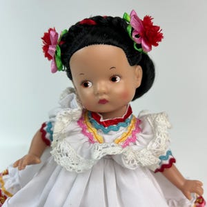 May include: A vintage doll with black hair styled in two buns, adorned with red and pink floral hair accessories. The doll wears a white dress with colorful embroidered details and a lace collar. The doll's skin tone is light brown.