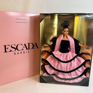 May include: A Limited Edition Escada Barbie doll in a display case. The doll wears a black dress with a pink skirt and a pink scarf. The box is pink with the Escada Barbie logo.