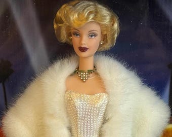 May include: A glamorous doll with blonde hair, wearing a white sequined gown and a fluffy white faux fur stole. She has a green and gold necklace and earrings. The doll is posed in front of a backdrop with a Hollywood theme.
