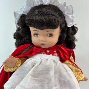 May include: A vintage doll with dark curly hair, wearing a red cape, white apron, and a white bonnet. The doll's dress has a floral pattern. The cape is trimmed with green and gold fabric. The doll has a serious expression.