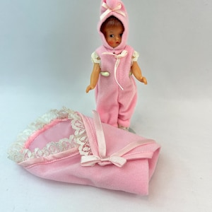 May include: A vintage doll wearing a pink hooded jumpsuit with a matching blanket. The doll has a fair complexion and is standing next to a pink blanket with white lace trim and a pink ribbon bow.