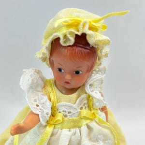 May include: A vintage doll with a painted face, wearing a yellow bonnet and dress with white lace trim. The doll's dress has yellow ribbon accents. The doll's hair is a reddish-brown color.