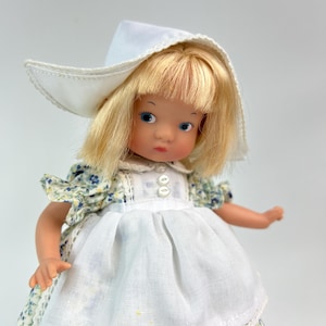 May include: A vintage doll with blonde hair and blue eyes, wearing a white bonnet and a floral print dress with a white apron. The dress features blue and yellow flowers. The doll is a collectible item.