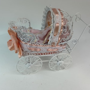 May include: A white metal baby carriage with intricate scrollwork and a pink and white interior. The carriage features a canopy, decorative trim, and large spoked wheels. A peach-colored fabric is draped on the side.