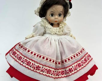 May include: A vintage doll with dark hair and a white lace bonnet. The doll is dressed in a white dress with a red and white embroidered skirt. A black bow with a red accent is on the doll's head.
