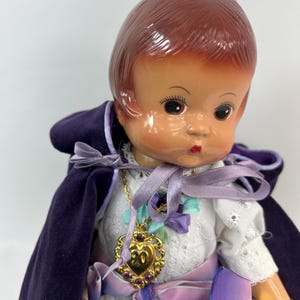 May include: A vintage doll with brown hair and a painted face, wearing a white dress with lace details and a purple velvet cape. The doll has a gold-colored heart-shaped pendant with purple accents and a purple ribbon sash.