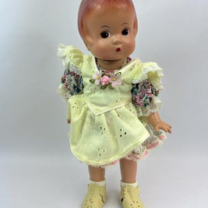 May include: A vintage doll with a reddish-brown molded hair, wearing a pale yellow dress with floral sleeves and a matching pair of shoes. The dress features eyelet detailing and is adorned with pink flower accents.