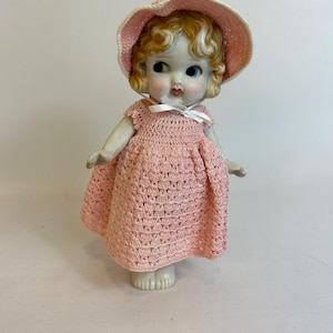 Puede incluir: Una mu&ntilde;eca de porcelana vintage con cabello rubio rizado, vestida con un vestido y sombrero de ganchillo rosa claro a juego. La mu&ntilde;eca tiene ojos azules y mejillas rosadas, y un lazo blanco en el escote. La mu&ntilde;eca est&aacute; sobre una superficie blanca.