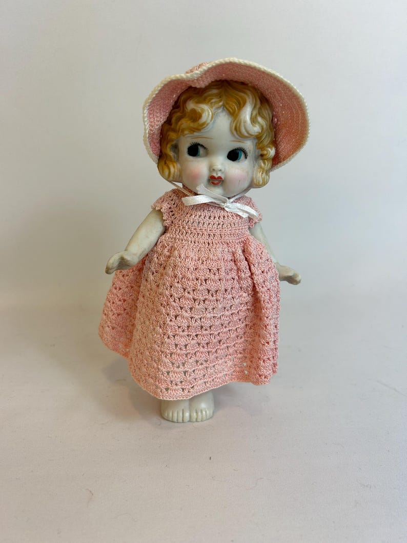 Puede incluir: Una mu&ntilde;eca de porcelana vintage con cabello rubio rizado, vestida con un vestido y sombrero de ganchillo rosa claro a juego. La mu&ntilde;eca tiene ojos azules y mejillas rosadas, y un lazo blanco en el escote. La mu&ntilde;eca est&aacute; sobre una superficie blanca.