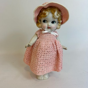 Puede incluir: Una mu&ntilde;eca de porcelana vintage con cabello rubio rizado, vestida con un vestido y sombrero rosa de ganchillo. La mu&ntilde;eca tiene ojos azules y mejillas rosadas, y una cinta blanca en el escote. La mu&ntilde;eca est&aacute; sobre una superficie blanca.