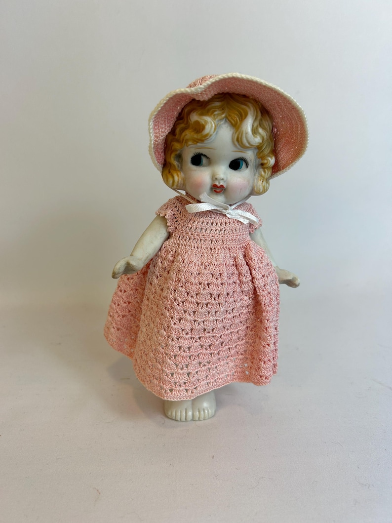 Puede incluir: Una mu&ntilde;eca de porcelana vintage con cabello rubio rizado, vestida con un vestido y sombrero rosa de ganchillo. La mu&ntilde;eca tiene ojos azules y mejillas rosadas, y una cinta blanca en el escote. La mu&ntilde;eca est&aacute; sobre una superficie blanca.