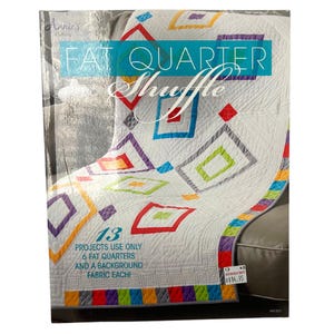 May include: A quilting pattern book titled Fat Quarter Shuffle by Annie's Quilting. The cover features a white quilt with colourful geometric square patterns in red, blue, green, orange, and purple, with text stating 13 projects use 6 fat quarters.