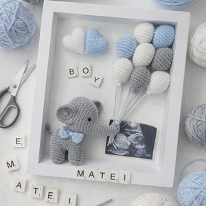 Op de afbeelding: Een witte ingelijste baby-aankondiging met een gehaakte grijze olifant die ballonvormige decoraties vasthoudt. De lijst bevat de naam "Matei" met lettertegels, een echo en bollen wol.