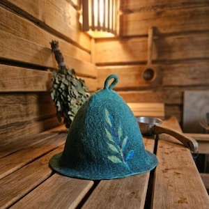 Può includere: Cappello da sauna color teal con un design a foglie. Il cappello è in feltro e ha una parte superiore arrotondata con un piccolo anello. È appoggiato su una panca di legno all'interno di una sauna. Sono visibili anche un fascio di rami frondosi e un mestolo di legno.