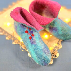 May include: A pair of handmade felt slippers. The slippers are primarily turquoise with a pink rolled cuff. The toe of each slipper is decorated with small red and purple dots. The slippers are designed for indoor use.