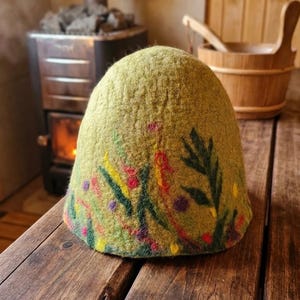 Può includere: Un cappello da sauna in lana infeltrita a forma di cupola di colore verde oliva chiaro. Il cappello presenta un motivo decorativo in lana agugliata con foglie verde scuro, sottili linee rosse e piccoli accenti circolari gialli e viola.
