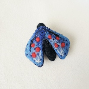 May include: A handmade felt moth brooch. The moth has blue wings with red spots and is embellished with small blue beads. The body is black. The brooch is a unique accessory.