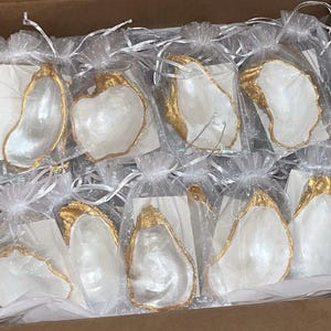 May include: A collection of white oyster shell dishes with gold accents, packaged in individual clear bags. The shells are arranged in a box, ready for gifting.