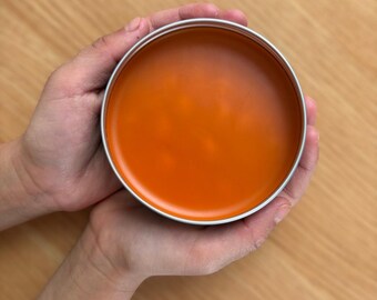 May include: A round, silver-colored metal tin containing a smooth, solid orange substance. The container has a simple, circular design with a metallic rim and a flat, uniform surface of the orange material filling the interior space.