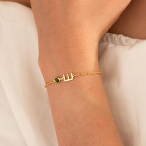 Op de afbeelding: Een verfijnde gouden kettingarmband met een kleine, ronde, levendig groene edelsteen en een enkele gouden letter E-bedel. De armband is gemaakt van een fijne, gepolijste gouden schakelketting die met een sluiting om de pols wordt bevestigd.