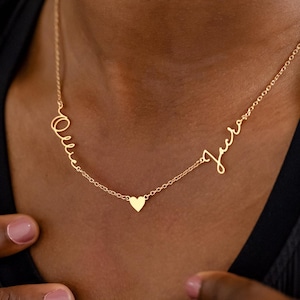 May include: Gold-coloured necklace featuring two names in a cursive script, with a small heart pendant. The necklace is worn against a black top.