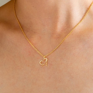 May include: A gold-coloured necklace with a heart-shaped pendant featuring the letter "A". The delicate chain and pendant are visible against a blue background.
