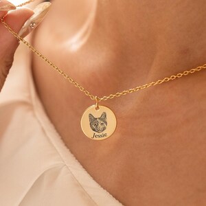 Puede incluir: Collar dorado con un colgante redondo que presenta un retrato detallado de un gato y el nombre "Jessie" grabado debajo. La cadena es delicada, y el colgante es sostenido por una mano con uñas cuidadas.