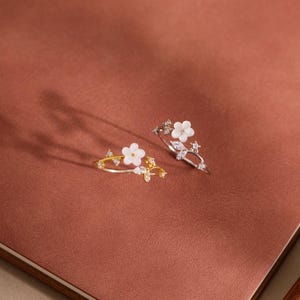 Peut inclure: Deux bagues délicates ornées d'un petit motif de fleur blanche en nacre, agrémentées de minuscules cristaux transparents sur un anneau métallique fin. Une bague est dorée et l'autre argentée, présentant toutes deux un design floral.