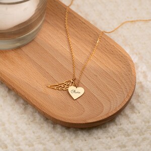 May include: A gold-colored necklace with a heart-shaped pendant and an angel wing charm. The heart pendant is engraved with the name "Olivia". The necklace is displayed on a wooden tray.