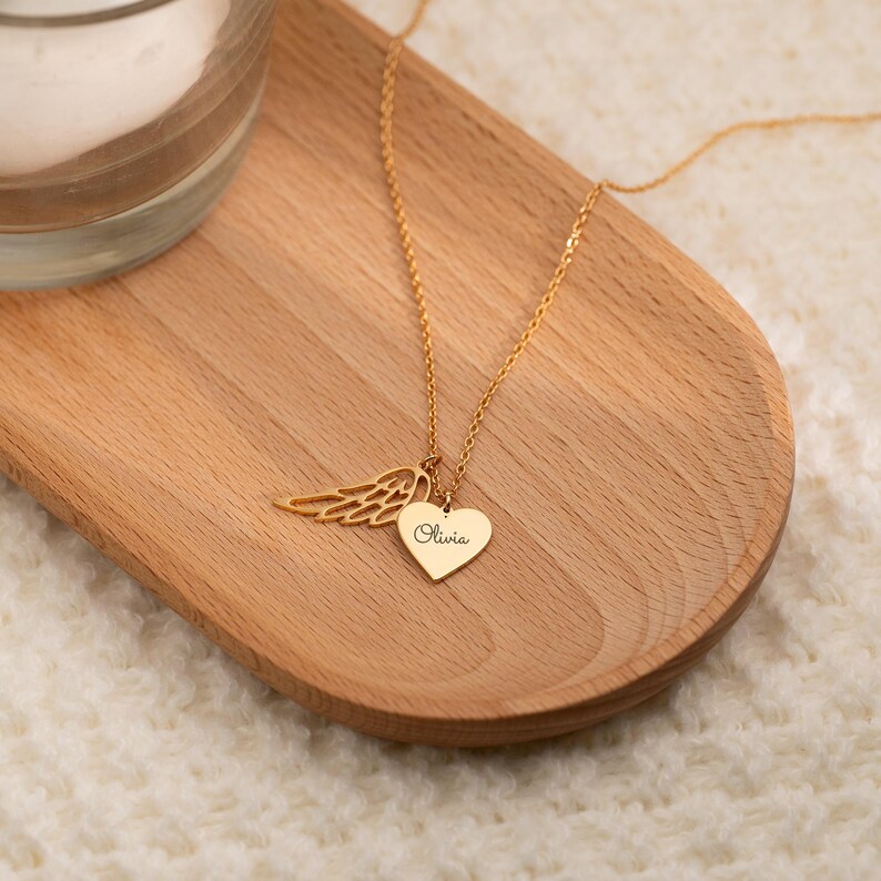 May include: A gold-colored necklace with a heart-shaped pendant and an angel wing charm. The heart pendant is engraved with the name "Olivia". The necklace is displayed on a wooden tray.