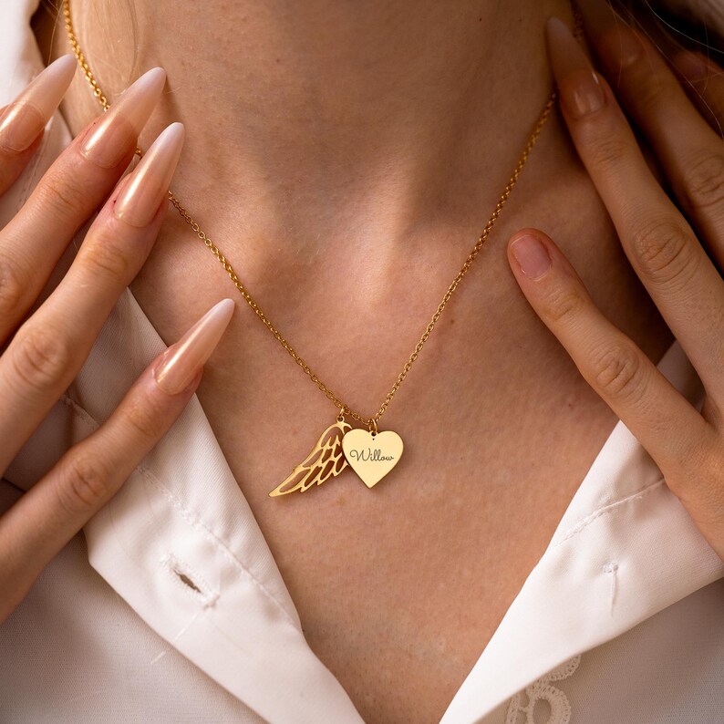 May include: Gold-colored necklace with a heart-shaped pendant and a wing charm. The heart pendant has the name "Willow" engraved on it. The necklace is worn over a white shirt.