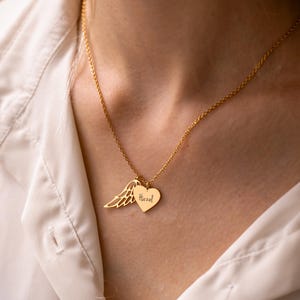 May include: A gold-colored necklace featuring two pendants. One pendant is a heart with the name "Hazel" engraved on it, and the other is a wing. The necklace is worn against a white shirt.