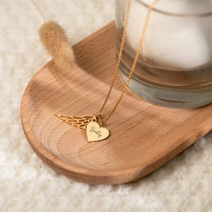 May include: A gold-colored necklace with a heart-shaped pendant and a wing charm. The heart pendant has the name "Bailey" engraved on it. The necklace is displayed on a wooden tray.