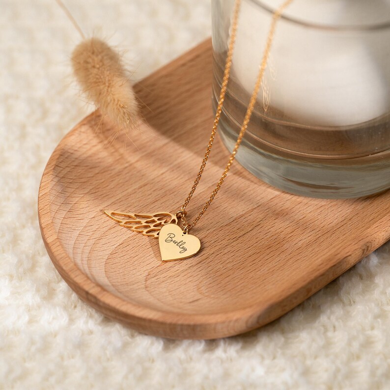 May include: A gold-colored necklace with a heart-shaped pendant and a wing charm. The heart pendant has the name "Bailey" engraved on it. The necklace is displayed on a wooden tray.