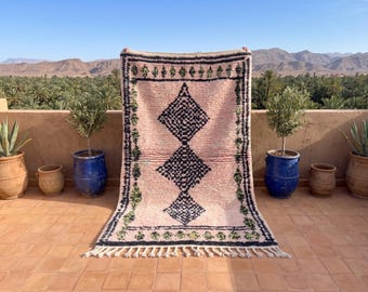 May include: A rectangular area rug featuring a soft pink background with a central vertical row of three large, dark navy blue diamond shapes. The rug is framed by a dark navy blue border with small green accents and finished with white fringe.