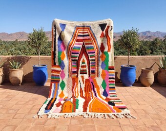 May include: A rectangular area rug with a cream-colored base featuring a vibrant, multi-colored geometric pattern. The design includes stacked horizontal stripes, diamond shapes, and abstract forms in orange, pink, blue, green, and black, with fringe.