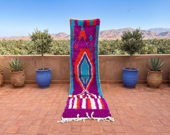 May include: A long, rectangular runner rug featuring a vibrant colour palette of magenta, bright orange, turquoise, and white. The design includes a large central diamond shape, geometric patterns, and a fringed edge at the bottom end.