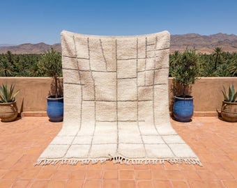May include: A large, rectangular, off-white wool rug featuring a grid pattern of thin, dark gray lines. The rug has a thick, shaggy pile texture and is finished with a short, knotted fringe along the bottom edge, creating a soft, textured surface.