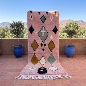 Puede incluir: Una alfombra rectangular de color rosa con un patrón geométrico de rombos en tonos azul oscuro, amarillo mostaza, blanco y azul claro. La alfombra tiene un pelo grueso y texturizado, rematada con flecos blancos en el borde inferior.