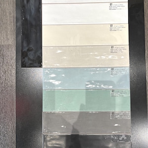 以下が含まれることがあります： 白、ベージュ、薄い青、緑、グレー、黒など、さまざまな色のセラミックタイルのサンプル8枚のディスプレイ。各タイルには、製品名、コレクション、サイズが記載されたラベルが付いています。