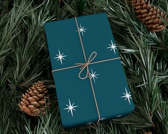 May include: A teal gift box wrapped with twine and adorned with white starburst patterns. The box is surrounded by pine needles and pine cones, creating a festive holiday theme.