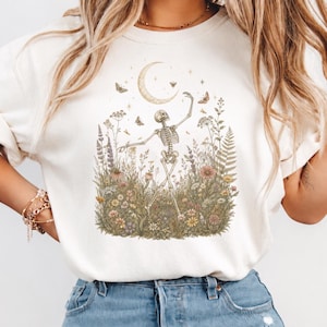 May include: A cream-colored short-sleeve t-shirt featuring a graphic print of a skeleton dancing in a field of wildflowers and greenery under a crescent moon. The illustration includes various small butterflies and stars surrounding the skeleton.