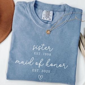May include: A blue Comfort Colors t-shirt with the words "sister EST. 1998 maid of honor EST. 2025" printed in white. A gold necklace with a pendant is on the shirt. A brown hat and dried plant are in the background.